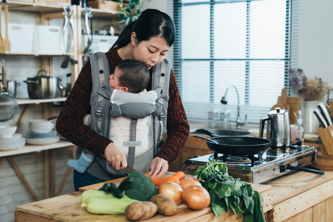 asian new mother is using a knife to cut vegetables with her newborn baby in the carrier in a modern bright kitchen at home.